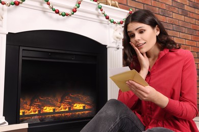 Photo of Emotional woman sitting near fireplace with greeting card indoors