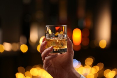 Man holding glass of whiskey with ice cubes against blurred lights, closeup.