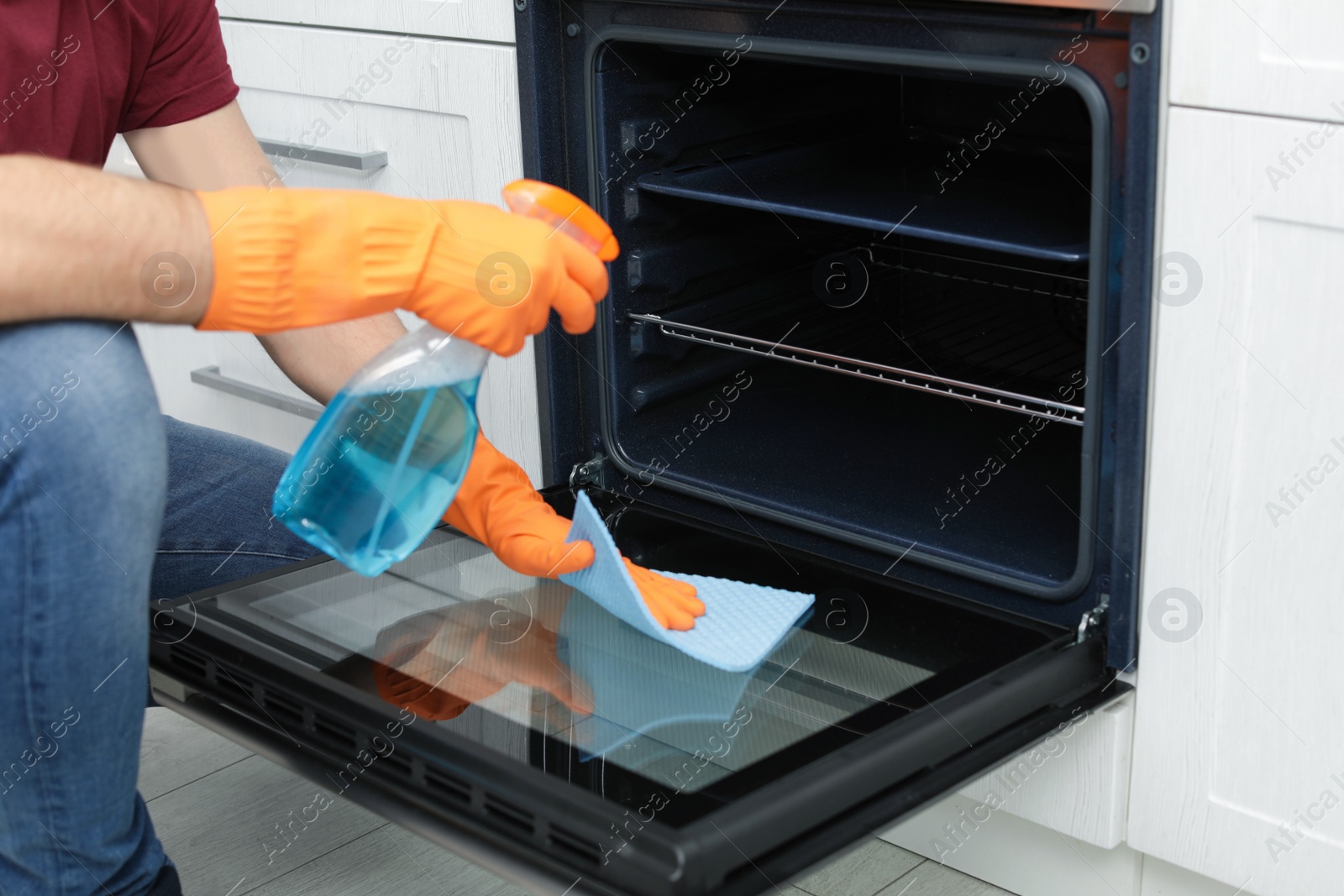 Photo of Young man cleaning oven with rag and detergent in kitchen, closeup