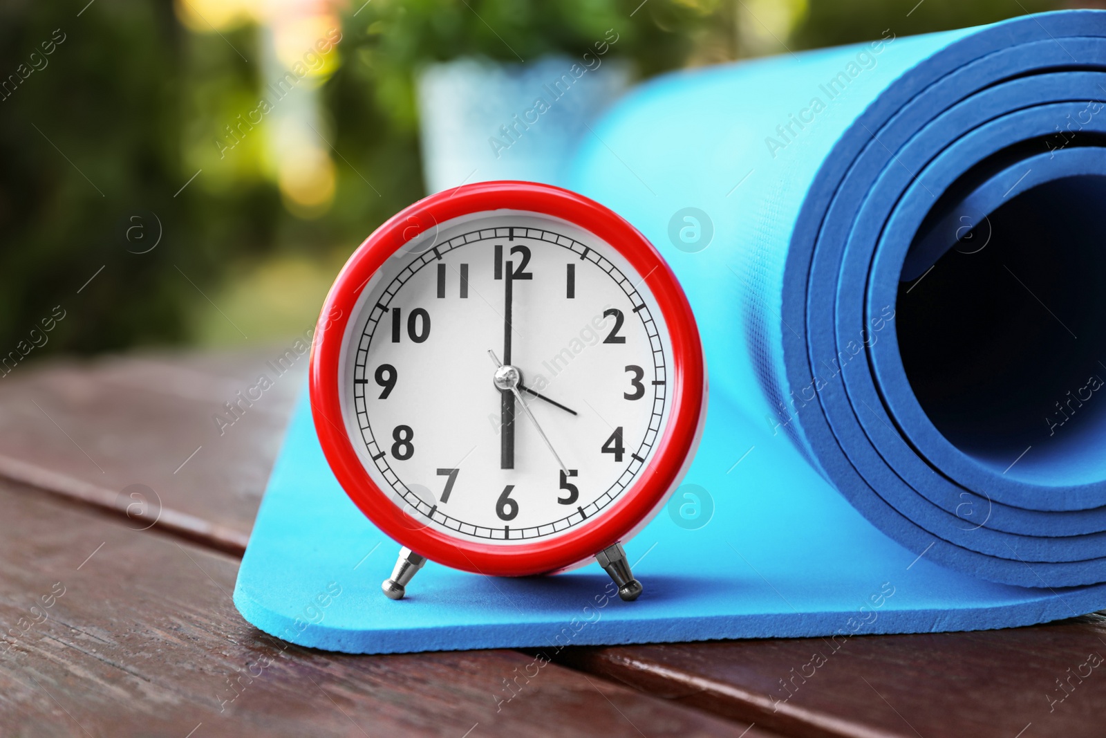 Photo of Alarm clock and fitness mat on wooden table outdoors, closeup. Morning exercise