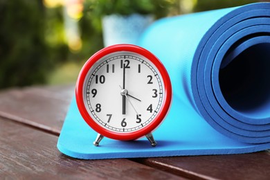 Photo of Alarm clock and fitness mat on wooden table outdoors, closeup. Morning exercise