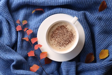 Cup of hot drink and leaves on blue knitted sweater, flat lay. Cozy autumn atmosphere