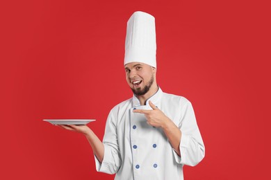Photo of Happy professional confectioner in uniform pointing at empty plate on red background