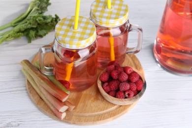 Photo of Mason jars of tasty rhubarb cocktail with raspberry and stalks on white wooden table