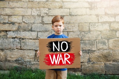 Photo of Sad boy holding poster with words No War against brick wall outdoors