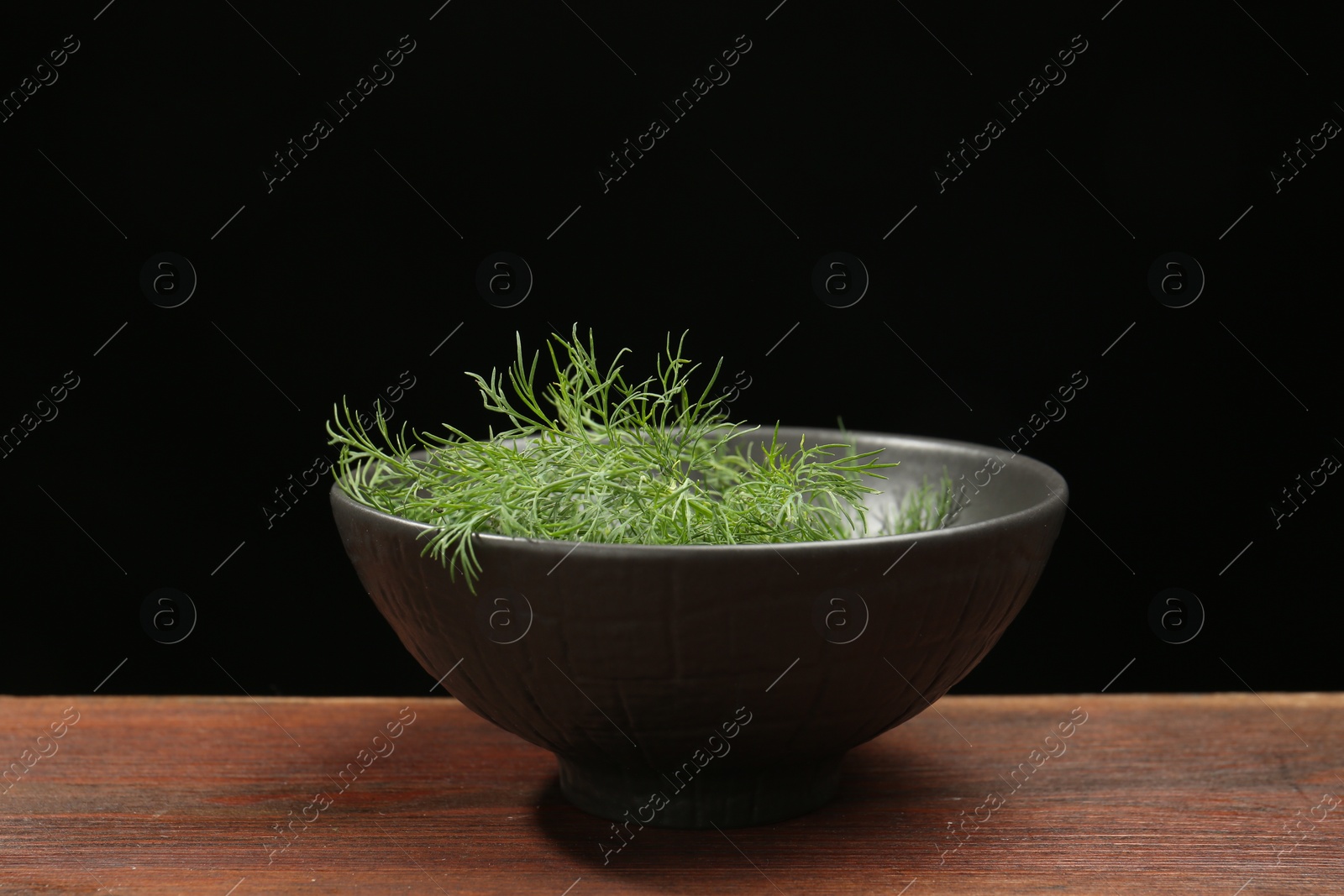 Photo of Bowl of fresh green dill on wooden table against black background