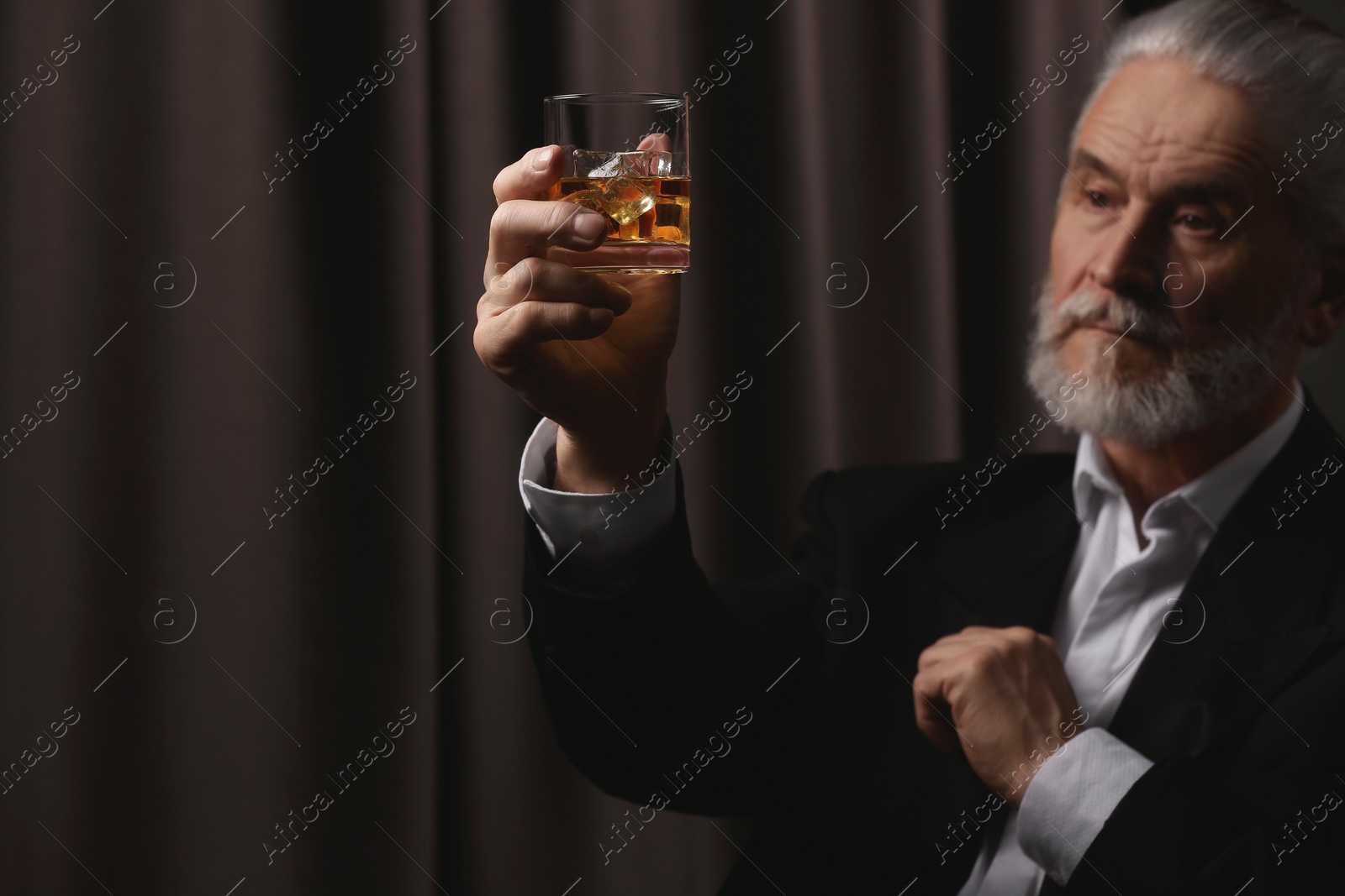 Photo of Senior man in suit holding glass of whiskey with ice cubes on brown background, selective focus