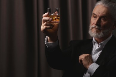 Senior man in suit holding glass of whiskey with ice cubes on brown background, selective focus