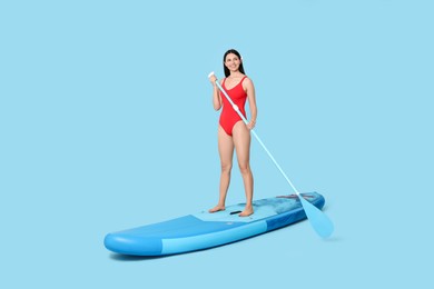 Happy woman with paddle on SUP board against light blue background, space for text