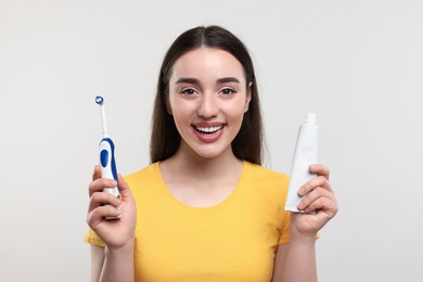 Happy young woman holding electric toothbrush and tube of toothpaste on white background