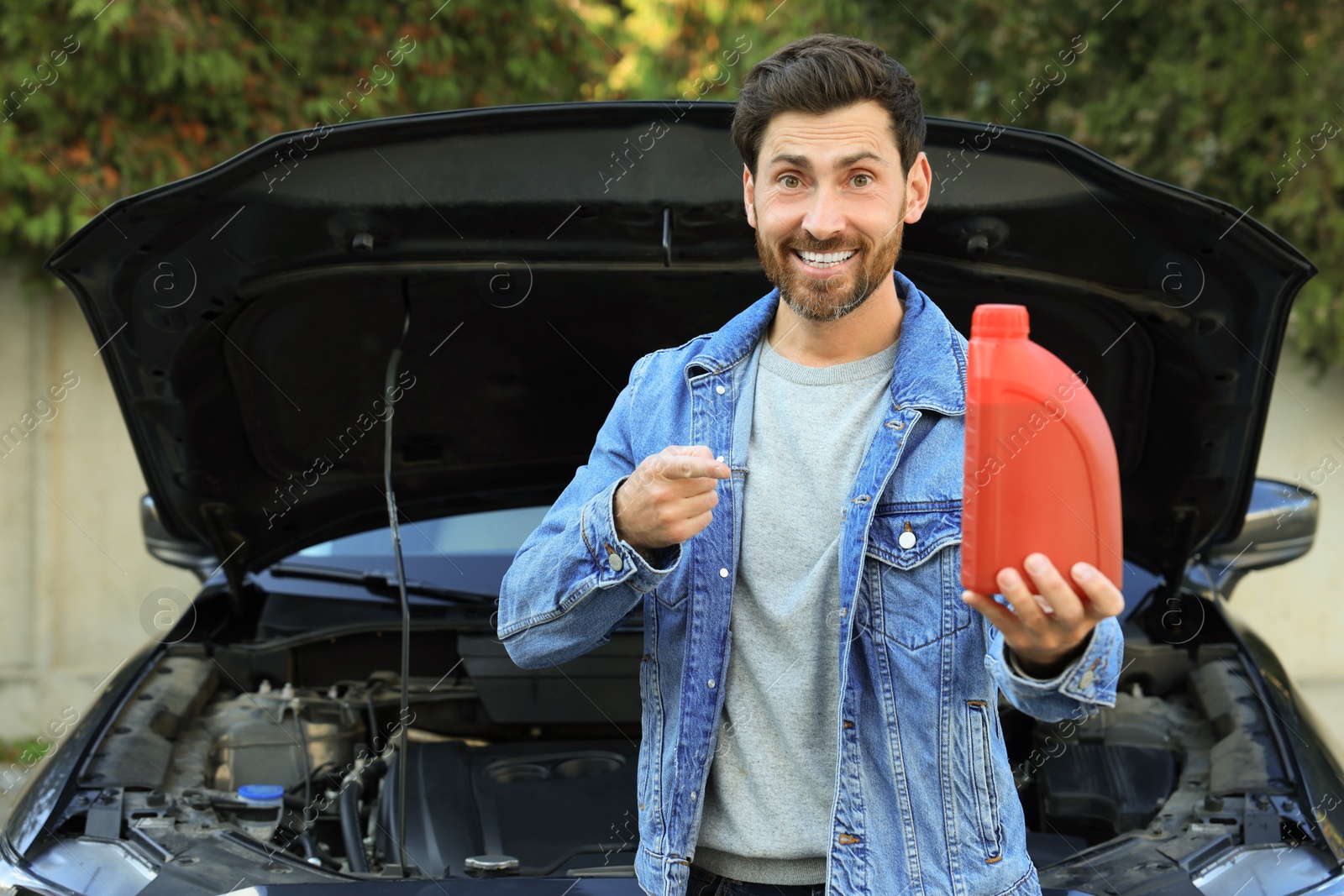 Photo of Smiling man pointing at red container of motor oil near car outdoors