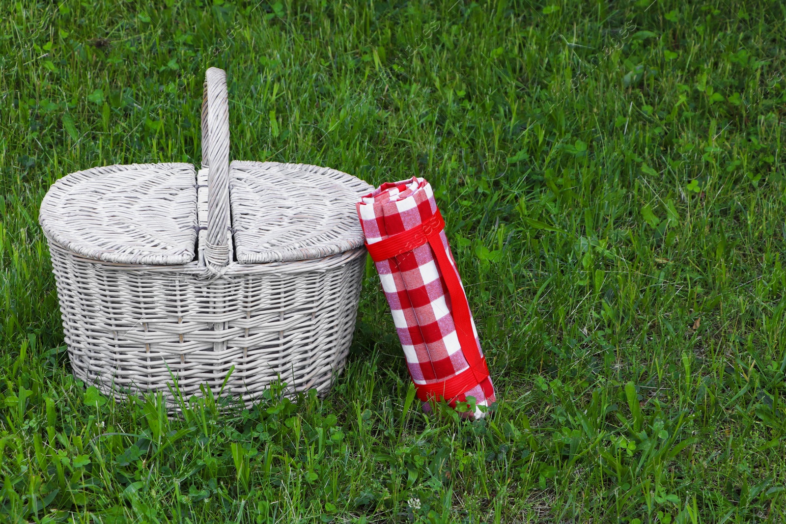 Photo of Rolled checkered tablecloth near picnic basket on green grass outdoors, space for text