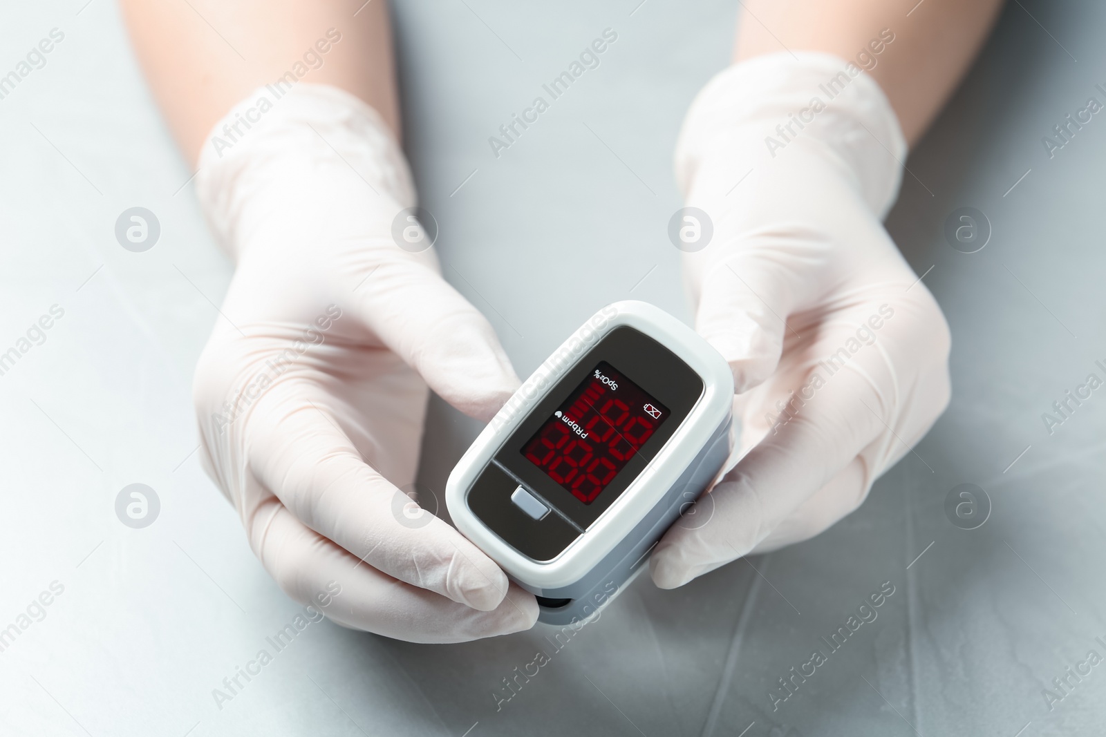 Photo of Doctor in gloves holding fingertip pulse oximeter at light grey table, closeup
