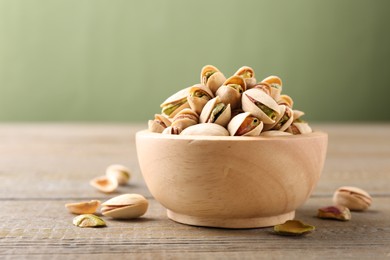 Tasty pistachios in bowl on wooden table against olive background, closeup