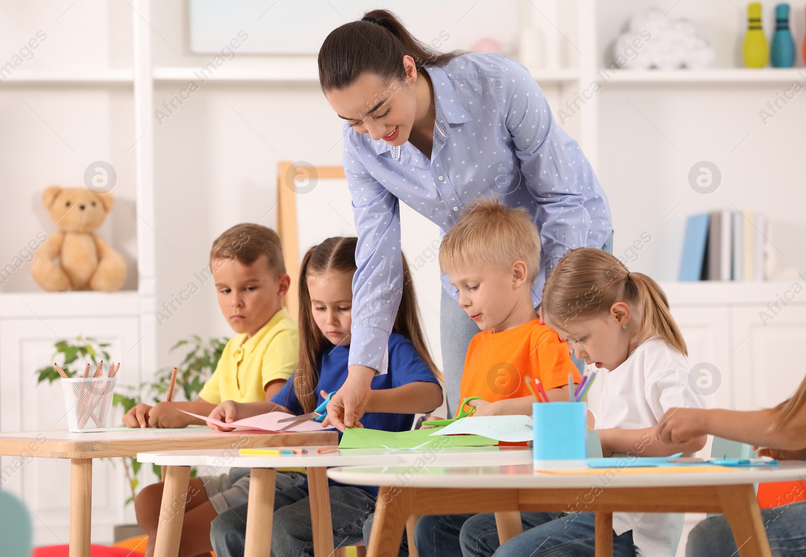 Photo of Nursery teacher with group of cute little children drawing and cutting paper at desks in kindergarten. Playtime activities