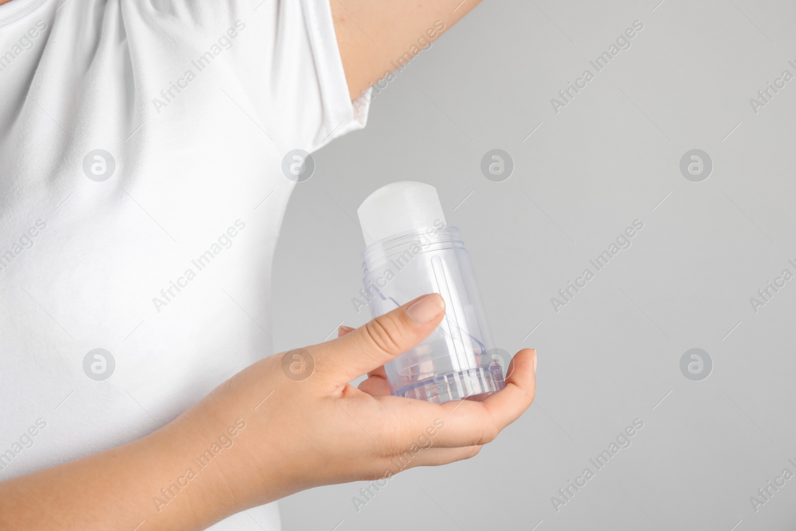 Photo of Young woman holding natural crystal alum deodorant near armpit on light grey background, closeup