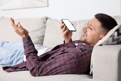 Photo of Handsome man recording voice message via smartphone on sofa at home