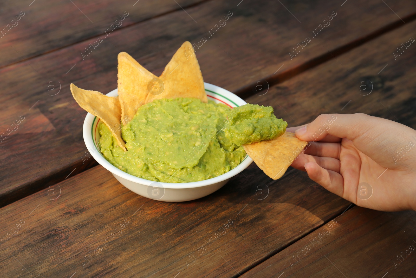 Photo of Woman holding nacho chip with delicious guacamole made of avocados at wooden table, closeup