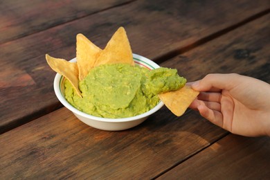 Photo of Woman holding nacho chip with delicious guacamole made of avocados at wooden table, closeup