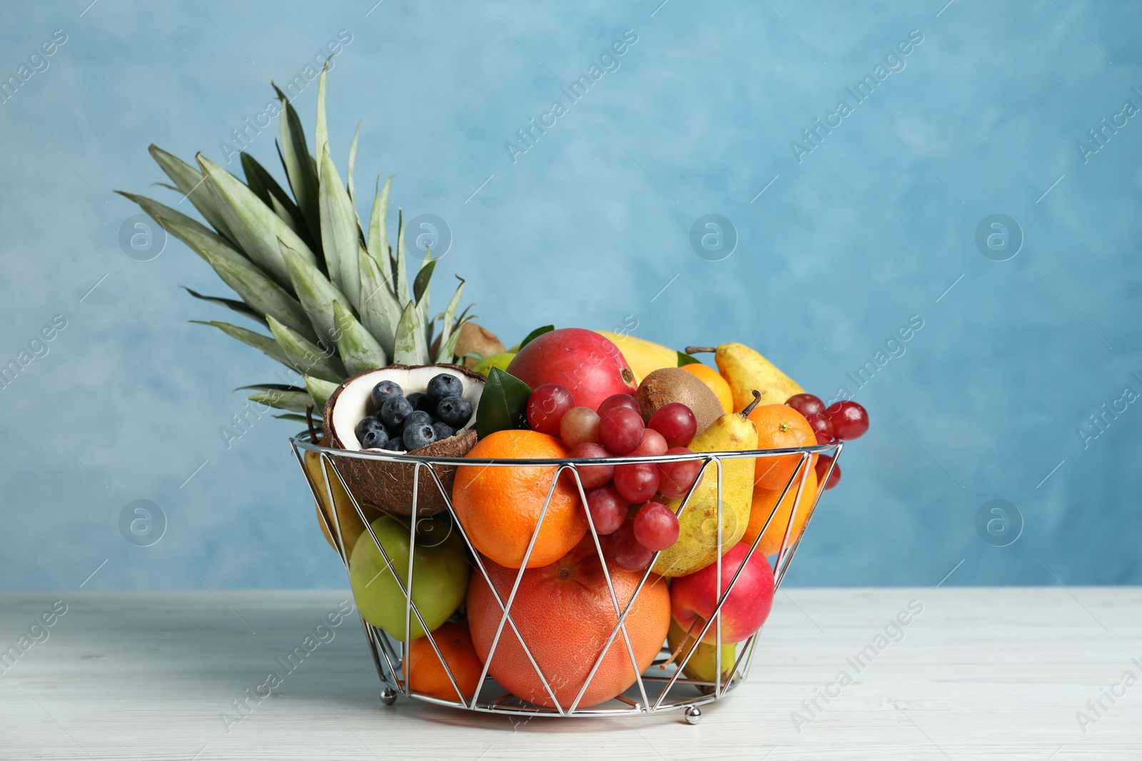 Photo of Assortment of fresh exotic fruits in metal basket on white wooden table against light blue background