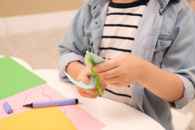 Little boy cutting color paper with scissors at table indoors, closeup