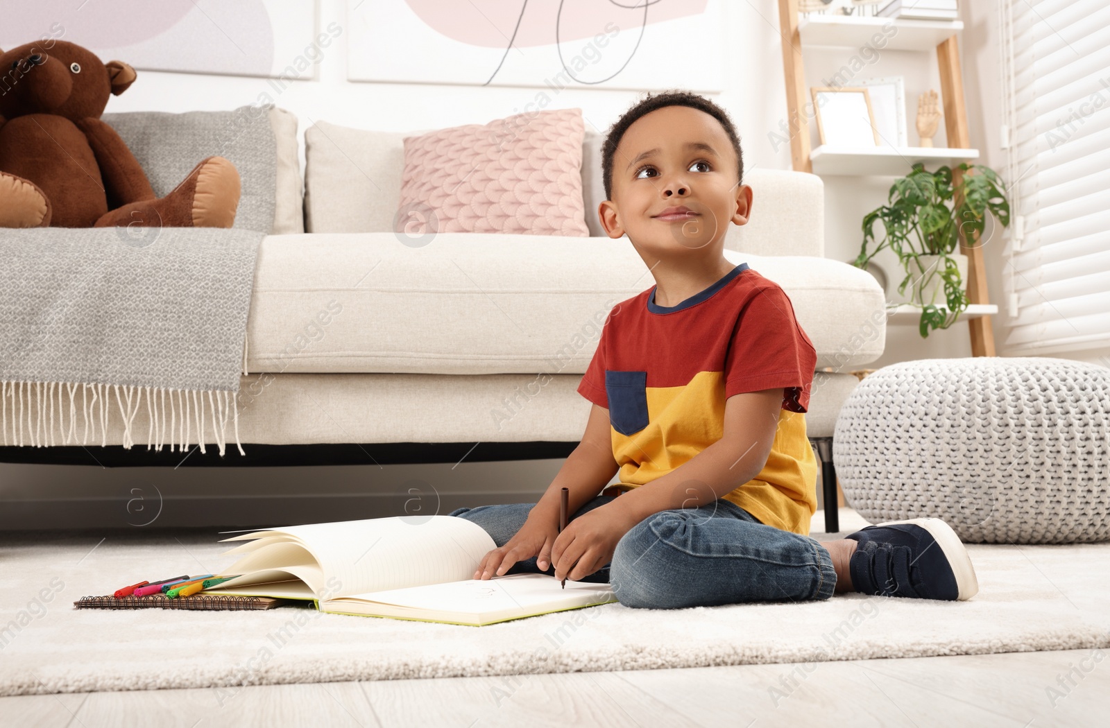 Photo of Cute African-American boy drawing in sketchbook with colorful markers on floor at home