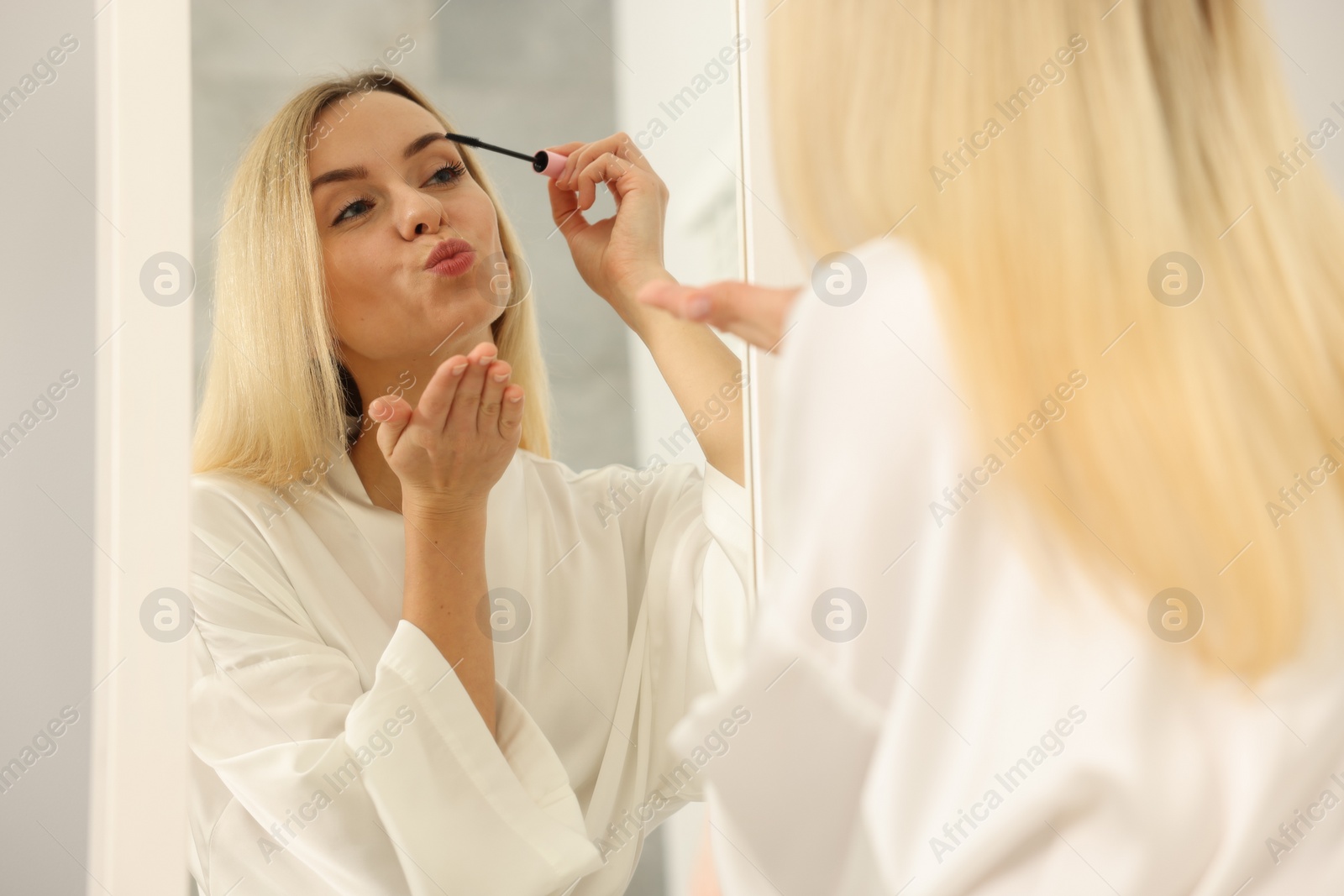 Photo of Beautiful woman blowing kiss while applying mascara near mirror in bathroom