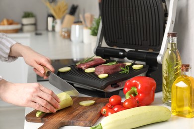 Photo of Woman cooking different products with electric grill at white wooden table in kitchen, closeup