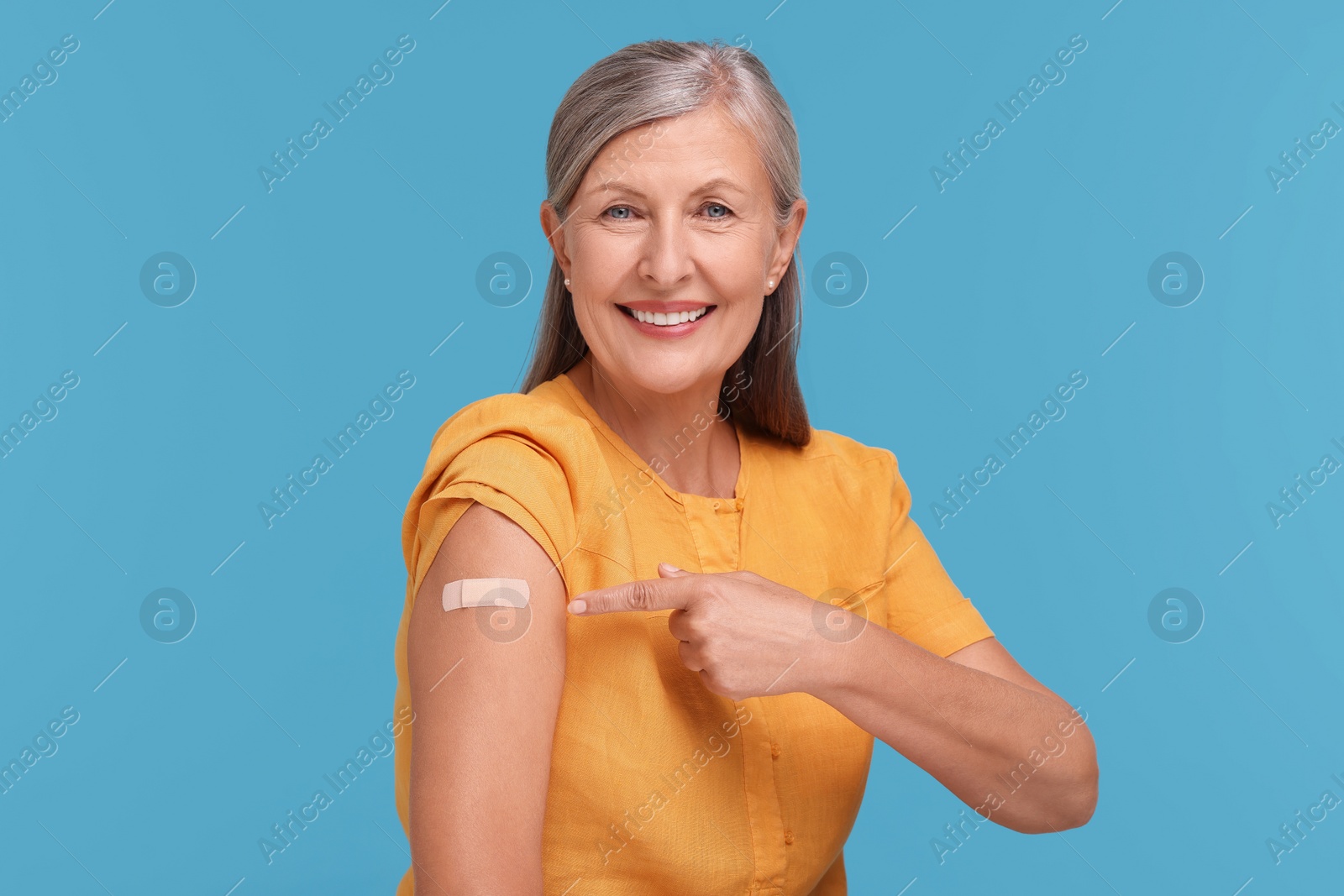 Photo of Senior woman pointing at adhesive bandage after vaccination on light blue background