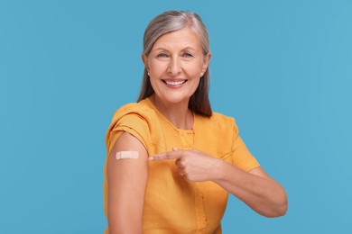 Photo of Senior woman pointing at adhesive bandage after vaccination on light blue background