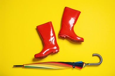 Red rubber boots near umbrella on yellow background, flat lay