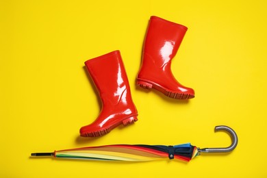 Photo of Red rubber boots near umbrella on yellow background, flat lay