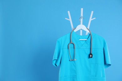 Photo of Turquoise medical uniform and stethoscope hanging on rack against light blue background. Space for text