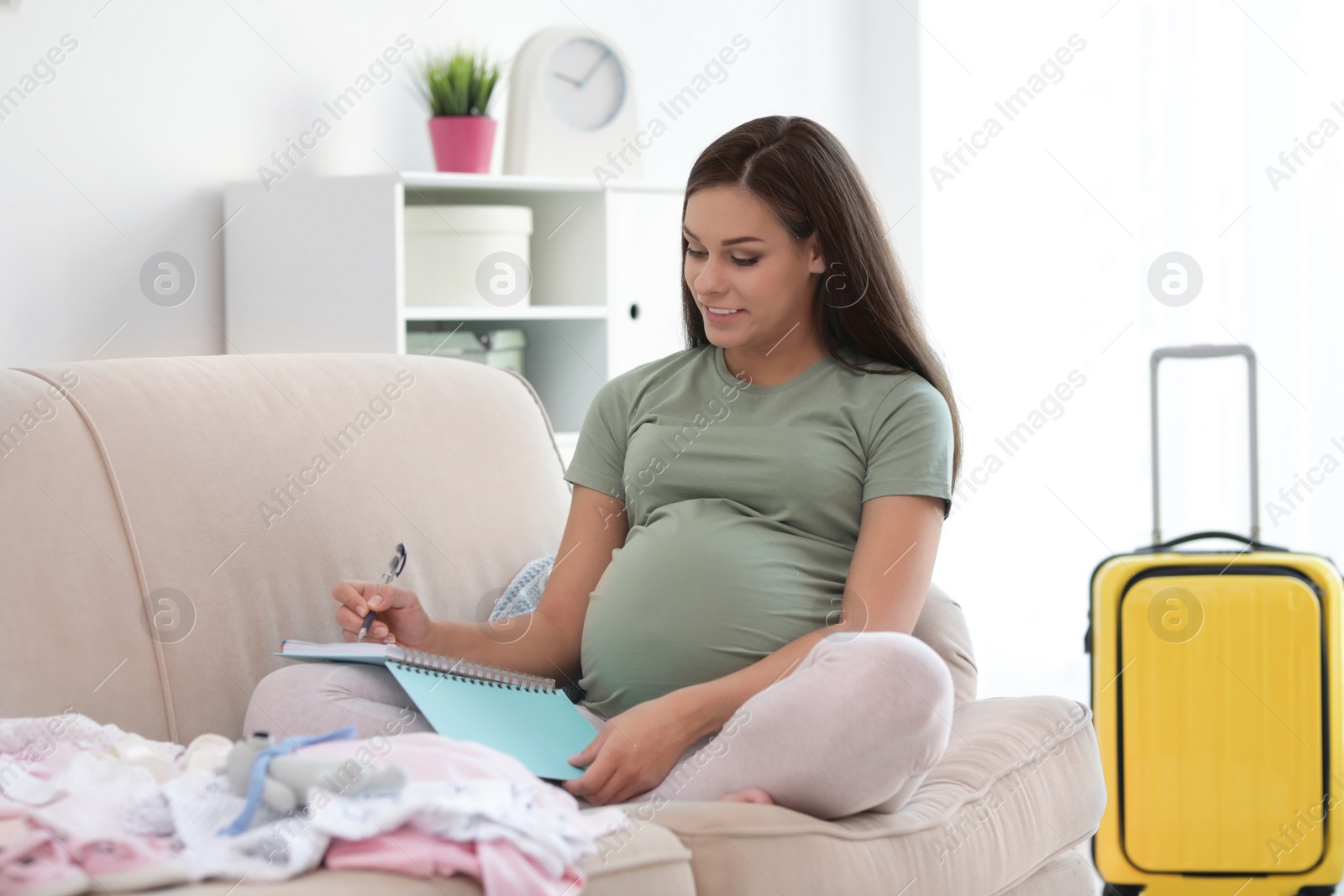 Photo of Pregnant woman making list while packing suitcase for maternity hospital in living room