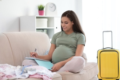 Pregnant woman making list while packing suitcase for maternity hospital in living room