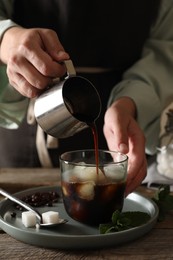 Woman pouring coffee into glass with ice cubes at wooden table, closeup