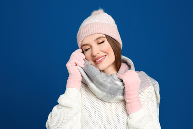 Photo of Young woman wearing warm sweater, gloves, scarf and hat on blue background. Winter season