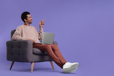 Photo of Happy man with laptop sitting in armchair and pointing at something on purple background. Space for text