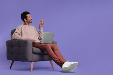 Happy man with laptop sitting in armchair and pointing at something on purple background. Space for text