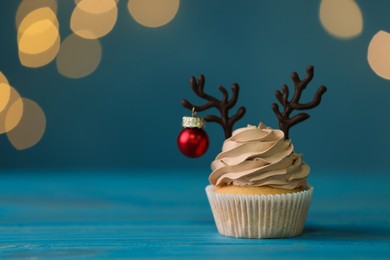 Photo of Tasty Christmas cupcake with chocolate reindeer antlers and bauble on blue wooden table against blurred festive lights, space for text