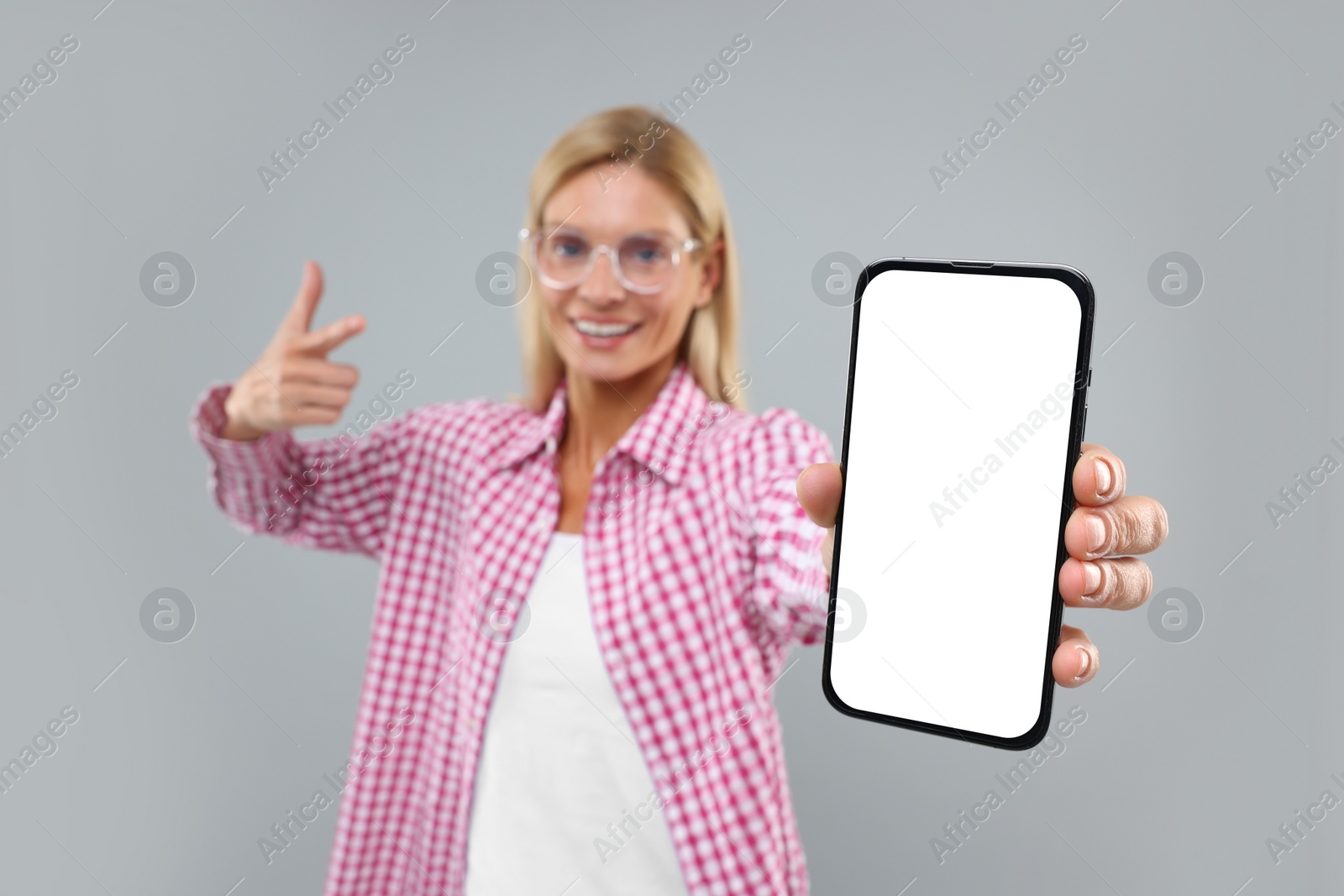 Photo of Happy woman holding smartphone and pointing at blank screen on grey background, selective focus