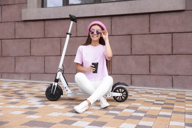 Photo of Young woman with cup of coffee sitting on electric kick scooter outdoors