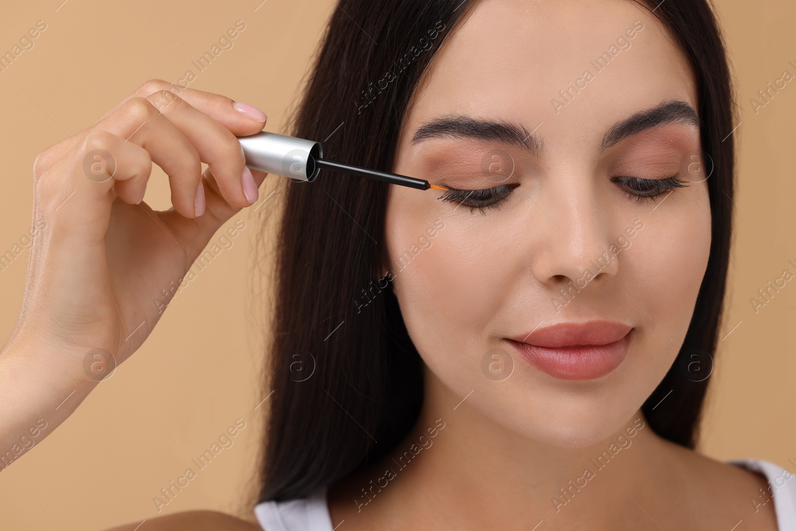 Photo of Beautiful woman applying serum onto her eyelashes on beige background. Cosmetic product