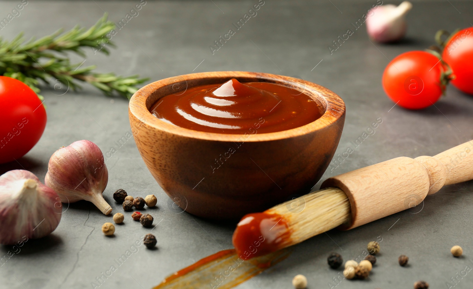 Photo of Tasty barbeque sauce in bowl, brush, garlic and peppercorns on grey table, closeup