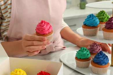 Woman putting delicious colorful cupcakes on dessert stand at white table indoors, closeup