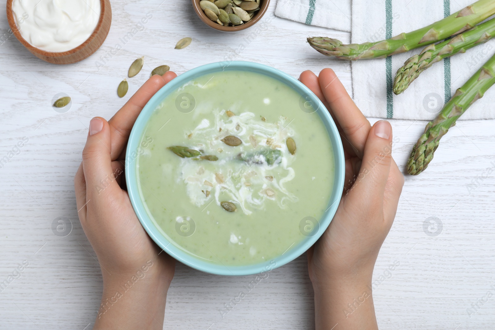 Photo of Woman with bowl of delicious asparagus soup at white wooden table, top view