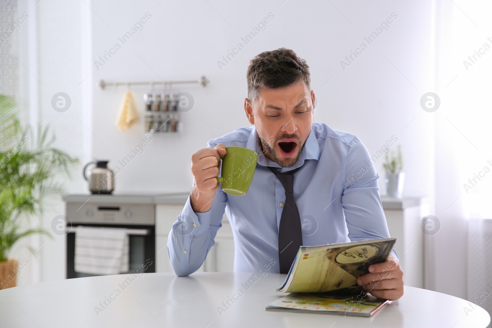 Photo of Sleepy man with cup of drink reading magazine at home in morning