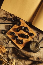 Many black rune stones, old book and dried plants on wooden table, flat lay