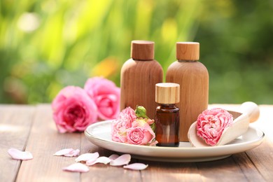 Bottles of rose essential oil and flowers on wooden table outdoors, space for text