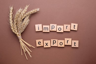 Words Import and Export made of wooden squares with ears of wheat on brown background, flat lay